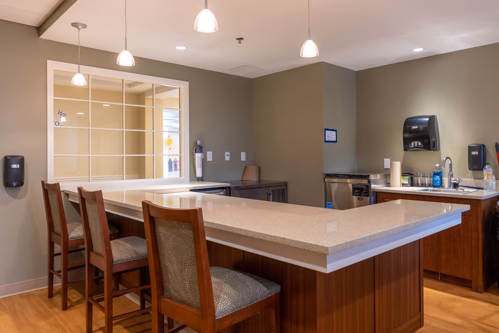 Interior view of a kitchen area with a large L-shaped countertop and four wooden chairs with patterned cushions. The kitchen features a sink, paper towel dispenser, soap dispenser, and various appliances. The walls are painted beige, and there are three pendant lights hanging from the ceiling.