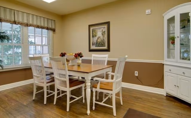 A dining room with a wooden table and six white chairs, one with armrests. The table has decorative bowls with flowers. There is a large window with blinds and a valance on the left wall, a framed picture on the beige wall, and a white china cabinet on the right side. The floor is wooden.