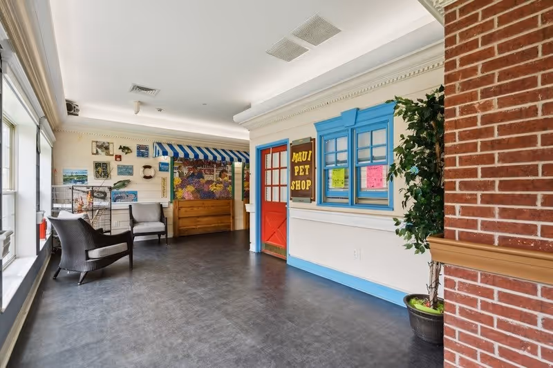 Interior hallway area with a pet shop storefront labeled 'Maui Pet Shop' featuring a red door and blue window frame. The hallway has large windows on the left side, two wicker chairs with cushions, various wall decorations, and a potted plant near a brick column on the right.