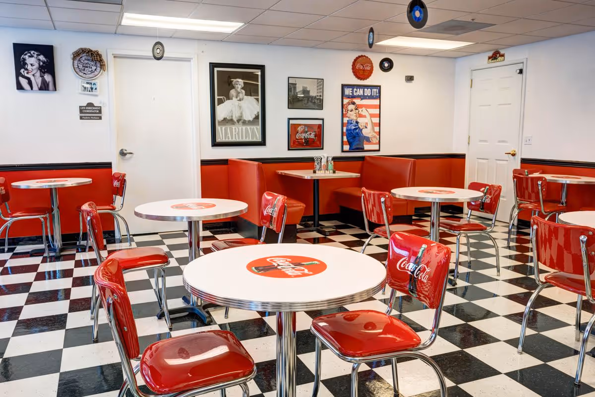 Retro-style dining room with red Coca-Cola chairs and round white tables on a black-and-white checkered floor.
