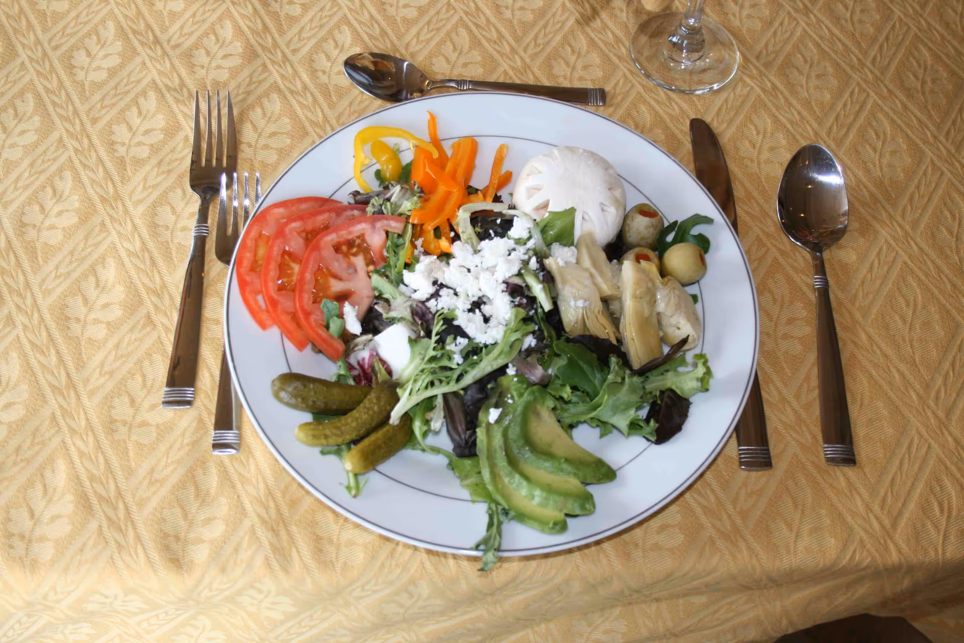 A plate of fresh salad on a yellow patterned tablecloth, featuring sliced tomatoes, pickles, avocado slices, mixed greens, shredded cheese, artichoke hearts, olives, and sliced bell peppers. The plate is accompanied by a fork, knife, and two spoons, with a glass of water partially visible at the top right.