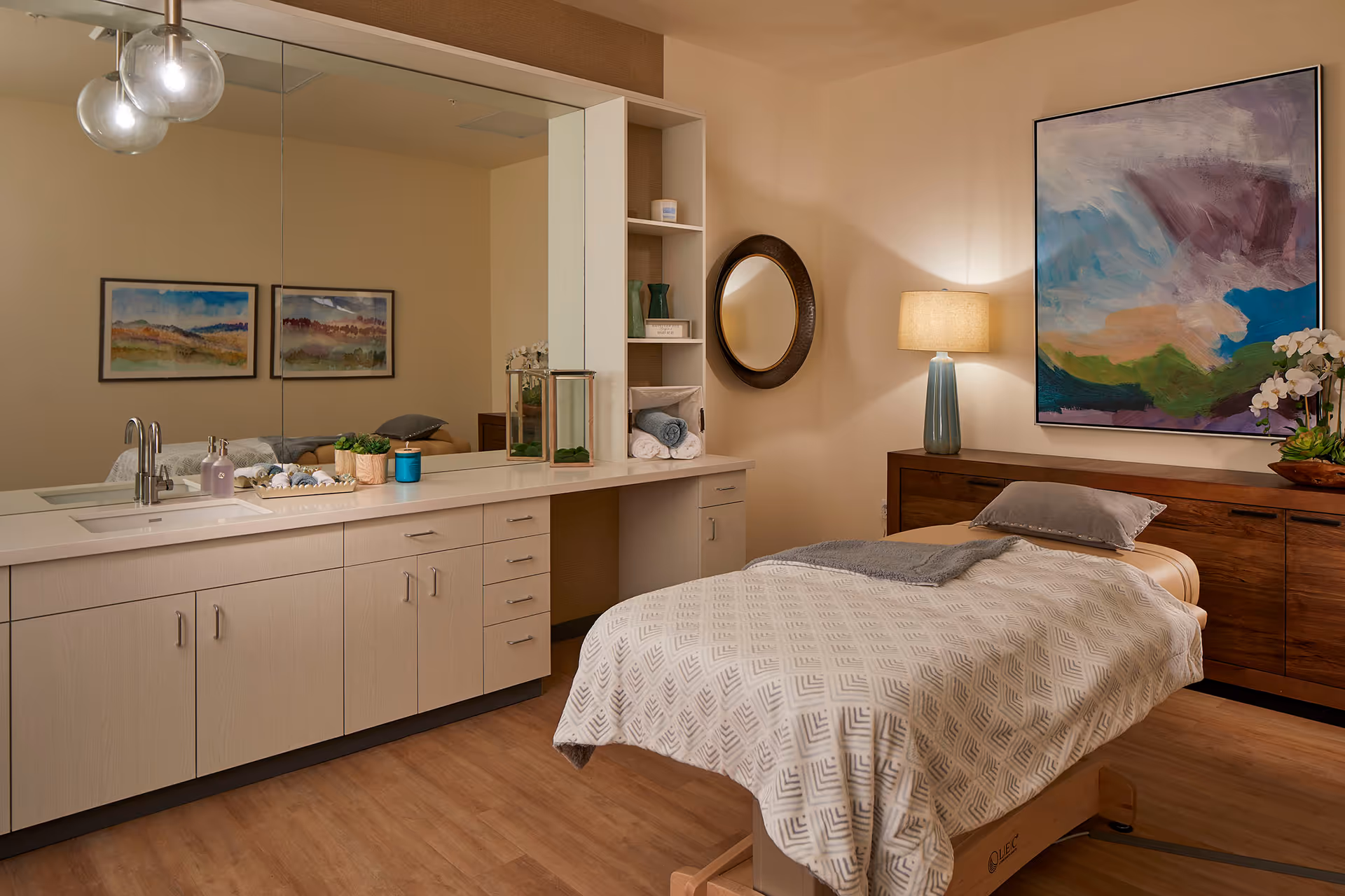 A cozy treatment or therapy room with a massage table covered in a patterned blanket and a pillow. The room features a large mirror above a countertop with a sink, decorative plants, and rolled towels. There is a round wall mirror, a table lamp with a blue base, and colorful abstract artwork on the wall. The floor is wooden, and the overall ambiance is warm and inviting.