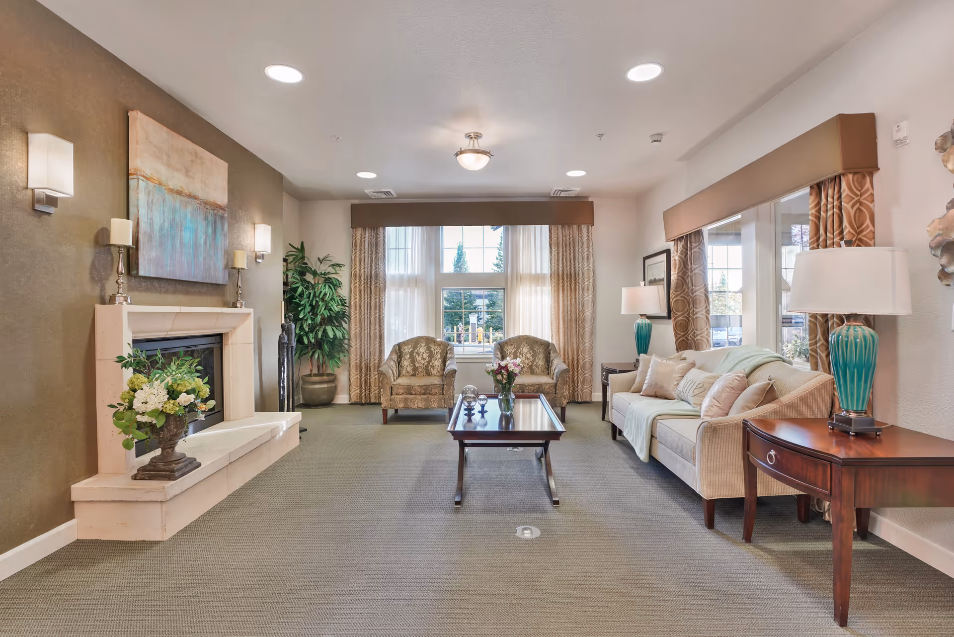 A well-lit living room with a beige sofa adorned with pillows and a throw blanket, two patterned armchairs near a large window with curtains, a wooden coffee table with a vase of flowers, a fireplace with decorative candles and a floral arrangement, and side tables with turquoise lamps. The room has a neutral color palette with green carpet and wall art above the fireplace.