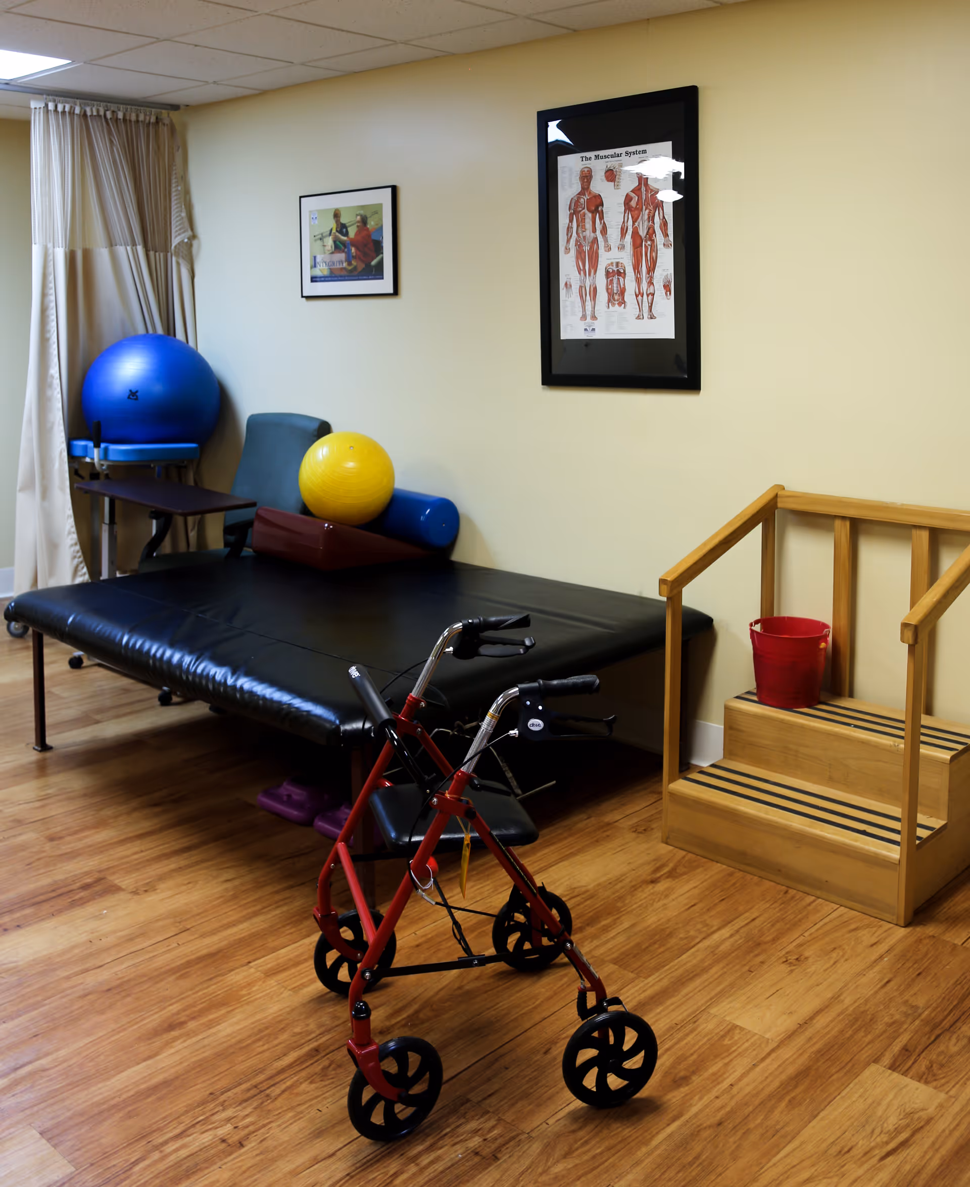 A senior rehabilitation therapy room with a red rolling walker in the foreground, a treatment mat, exercise balls, wooden therapy steps, and an anatomical poster on the wall.