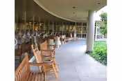 Curved outdoor covered walkway with wooden rocking chairs lined up along the glass wall of a building, with greenery visible on the right side.