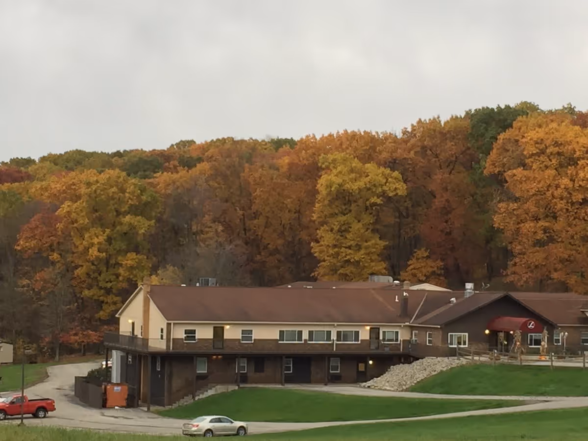 Front exterior of a two-story Elmcroft senior living building with a driveway, parked cars, and colorful autumn trees on the hillside behind it.