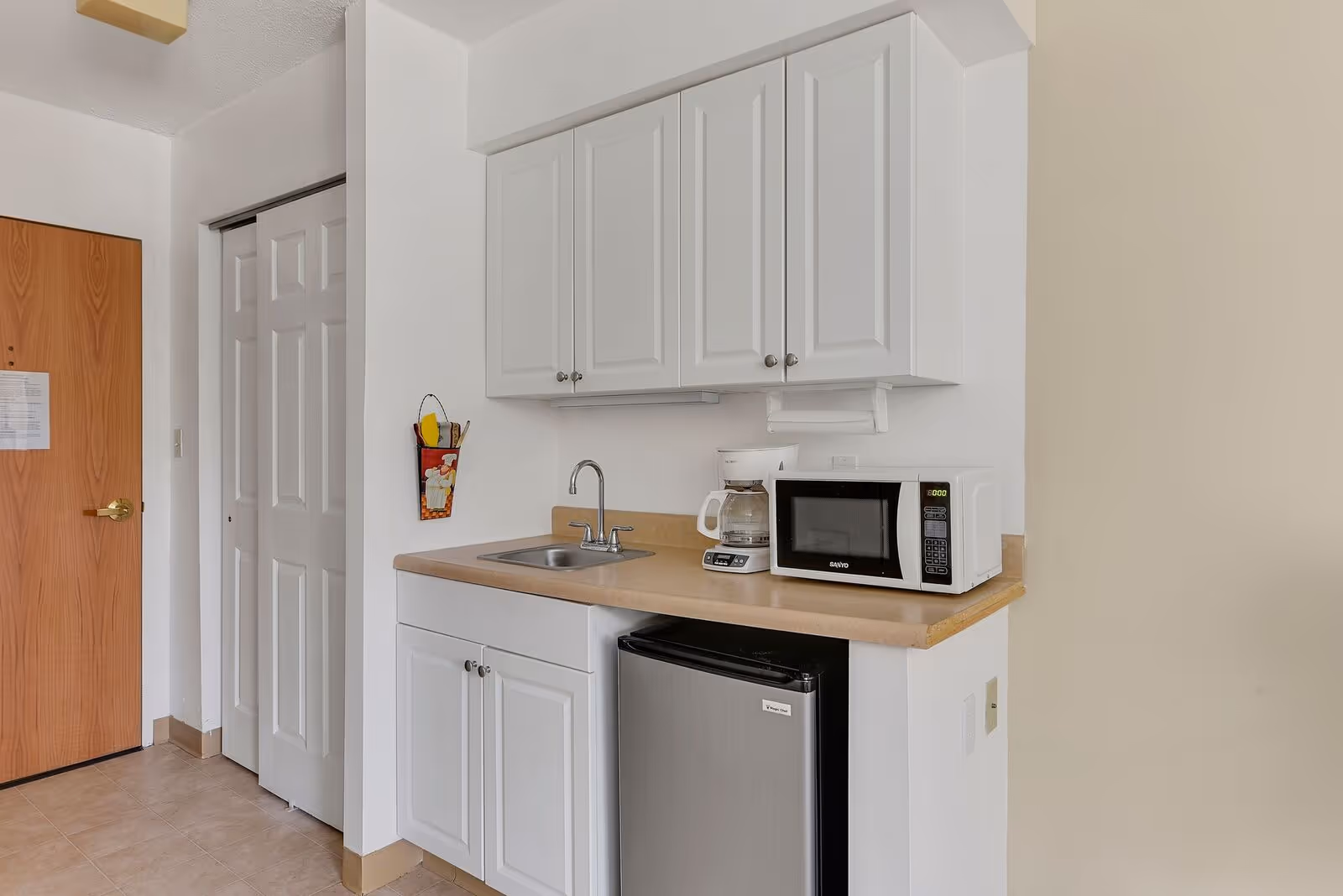 Small kitchenette area with white cabinets, a countertop with a sink, a coffee maker, a microwave, and a mini refrigerator. There is a wooden door and a white closet door in the background.