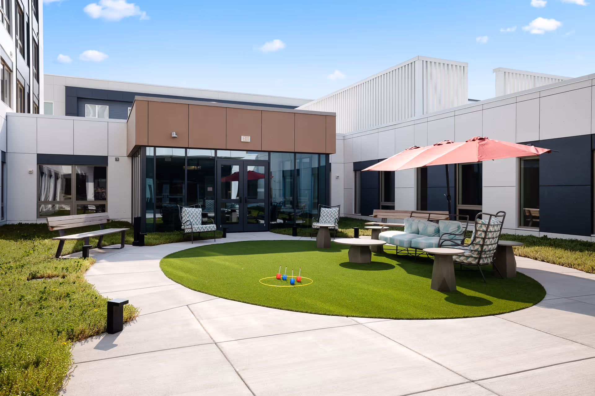 Outdoor courtyard area at Revelle King of Prussia featuring a circular patch of artificial grass with a ring toss game set up. Surrounding the grass are cushioned chairs, a sofa under a large red umbrella, wooden benches, and a concrete walkway. The courtyard is enclosed by modern building walls with large windows and glass doors.