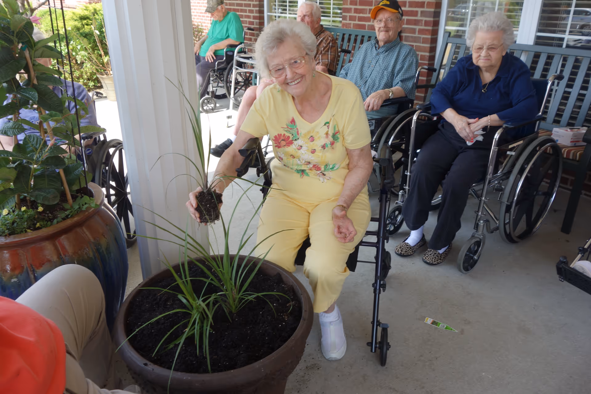 A group of elderly people, most in wheelchairs, are sitting on a covered patio. One elderly woman in a yellow outfit is smiling and holding a small plant, about to place it in a large planter filled with soil. There are other potted plants nearby and a brick wall in the background.