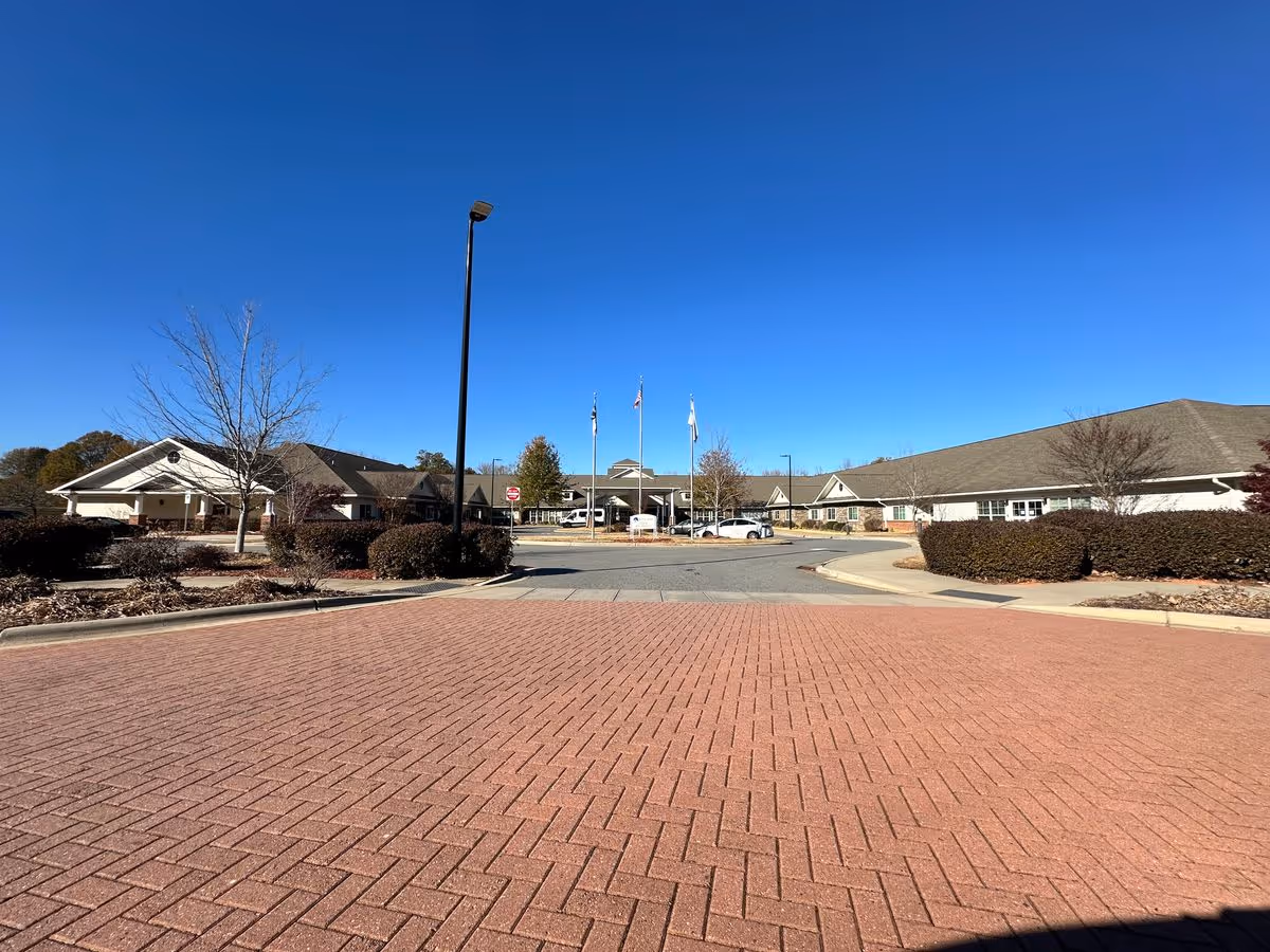 Front entrance of a single-story senior living facility with a circular driveway, flagpoles, and landscaped shrubs under a clear blue sky.