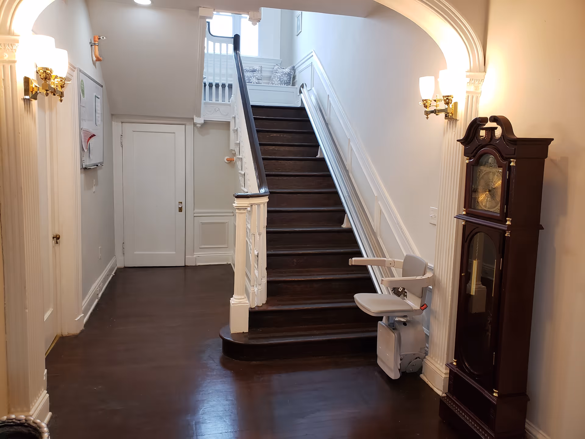 Interior entry hall with a dark wooden staircase fitted with a stairlift, a grandfather clock, and wall sconces under an arched doorway.