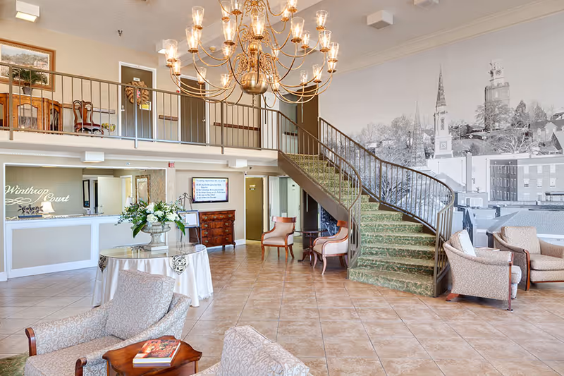 Spacious senior living lobby with seating areas, a reception desk, a large chandelier, and a curved carpeted staircase against a muraled wall.