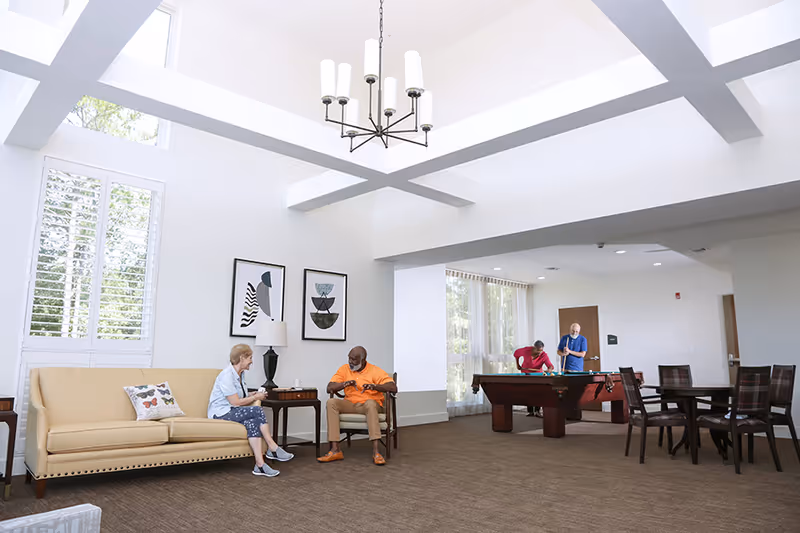 A bright and spacious common area in an assisted living facility with high ceilings and large windows. Two elderly people are seated and conversing near a beige couch and a side table with a lamp. In the background, two other elderly individuals are playing pool. There are several tables and chairs arranged around the room.