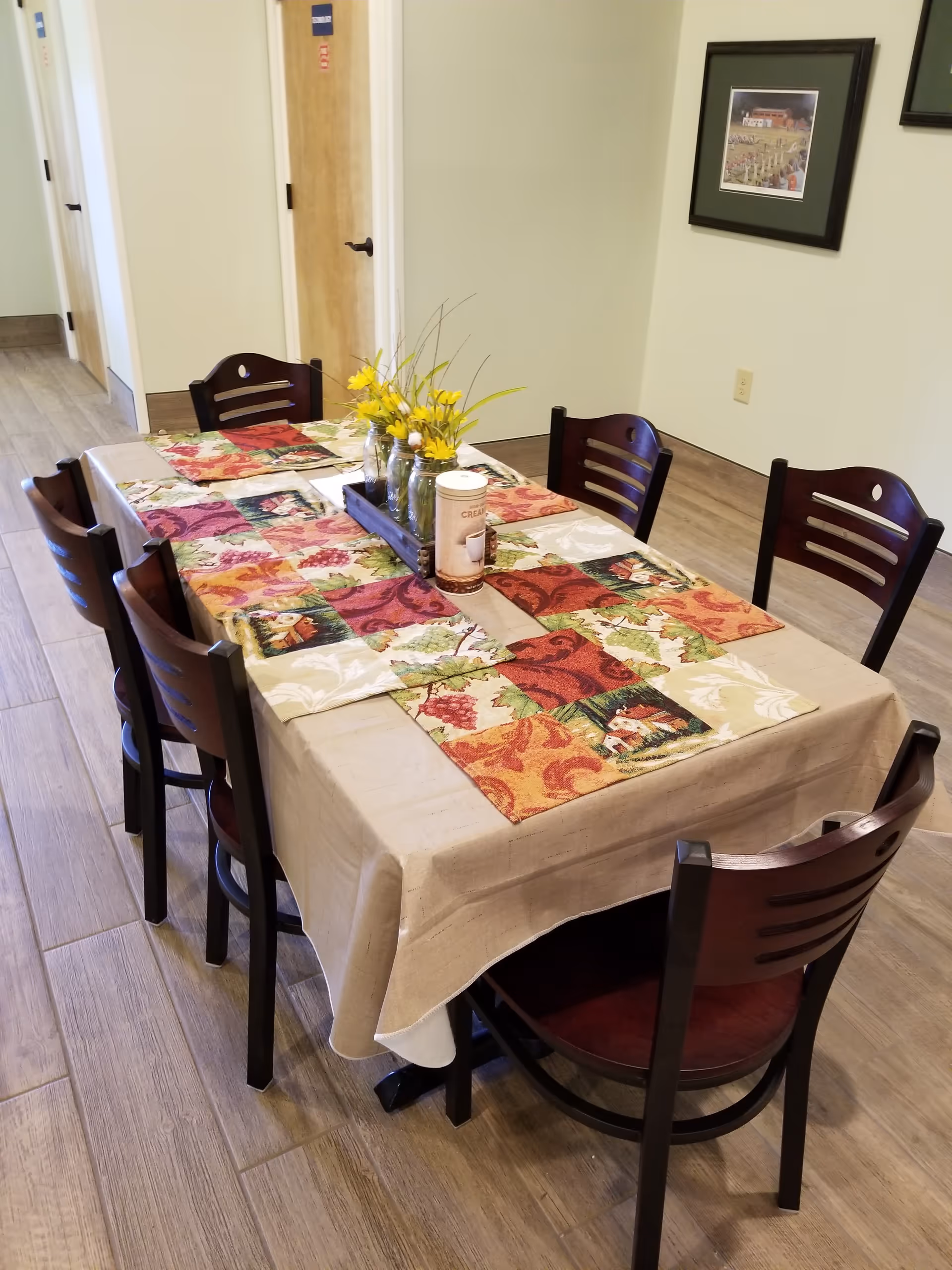 A dining table set with six dark wooden chairs in a room with light-colored walls and wood flooring. The table is covered with a beige tablecloth and colorful placemats featuring autumn-themed patterns. A centerpiece with yellow flowers and a container labeled 'CRISP' is placed on the table. A framed picture hangs on the wall in the background.