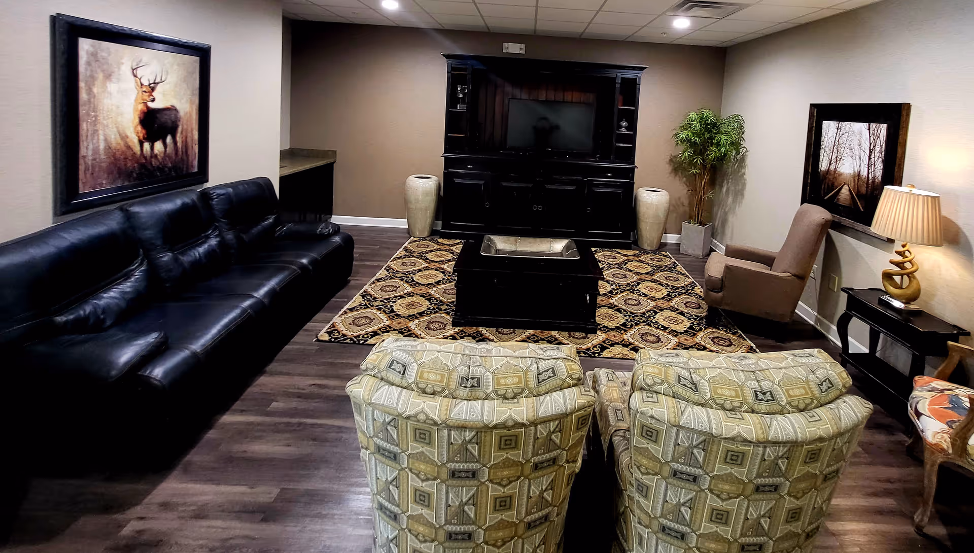 A cozy living room with a black leather sofa on the left, two patterned armchairs facing a dark wood coffee table and entertainment center with a TV. The room has a patterned area rug, a potted plant in the corner, a side table with a lamp, and framed artwork on the walls.