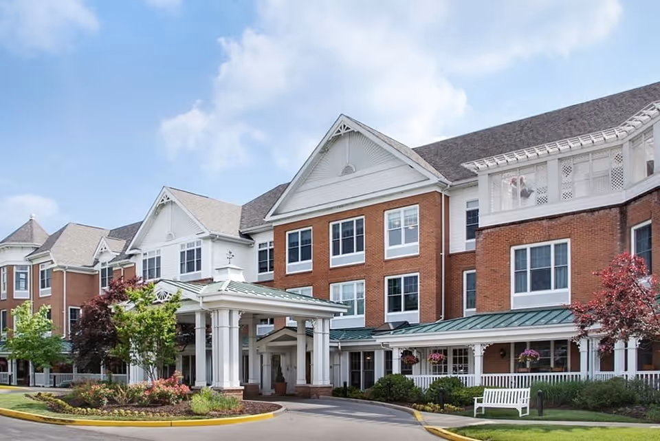Exterior view of a large senior living facility building with red brick and white siding, multiple windows, a covered entrance with white columns, landscaped greenery, and a white bench near the entrance under a partly cloudy sky.