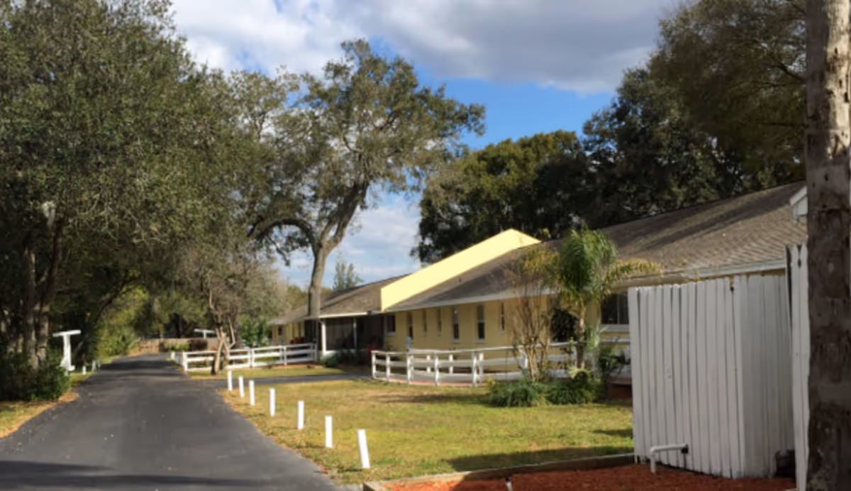 A paved driveway lined with white posts leads past a single-story yellow building with a sloped roof, surrounded by green trees and a white fence under a partly cloudy sky.