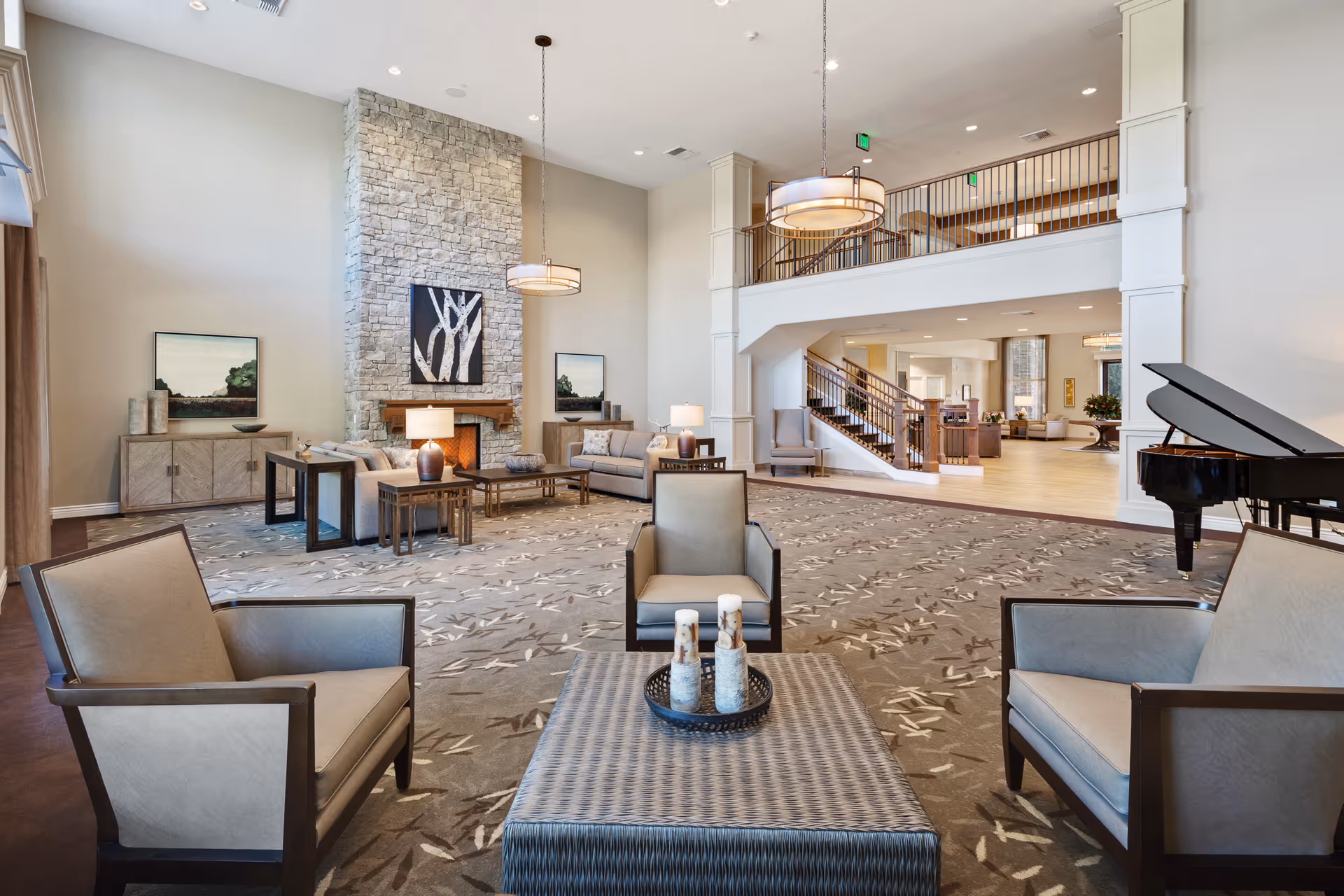 Spacious and well-lit senior living facility common area with high ceilings, a stone fireplace, modern furniture including armchairs and sofas, a large patterned carpet, a grand piano, and a staircase leading to an upper level.