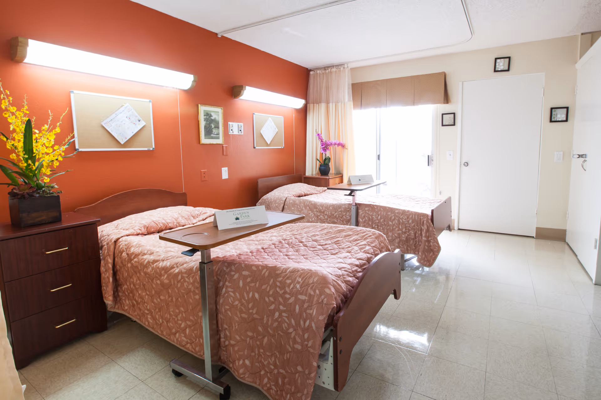 A bright and clean bedroom in a care facility with two single beds covered in matching pink patterned bedspreads. Each bed has an overbed table with a sign that reads 'Garden Park Care Center.' There is a wooden nightstand with a yellow flower arrangement between the beds. The walls are painted white and orange, with two wall-mounted lights above the beds. A window with sheer curtains and a brown valance lets in natural light. The floor is tiled and the room has a simple, tidy appearance.