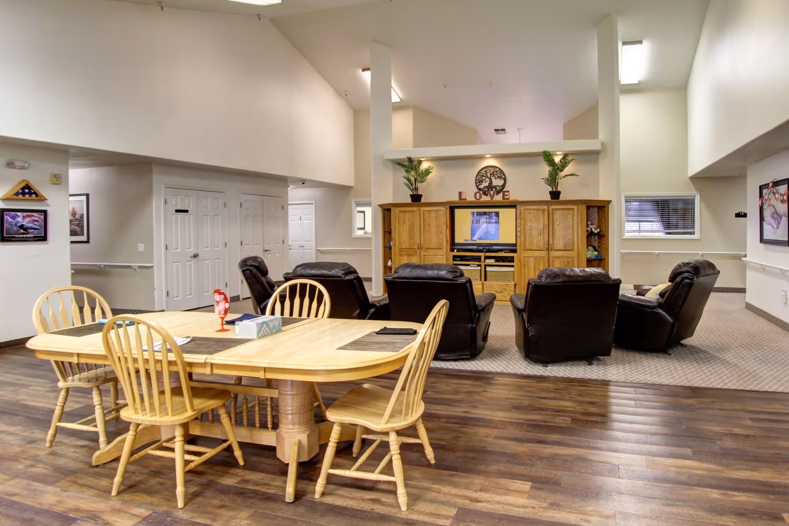 A senior living facility common area with a wooden dining table and four chairs in the foreground. Behind the table, there are five dark leather recliners arranged in a semicircle facing a wooden entertainment center with a TV. The entertainment center is decorated with two potted plants and a decorative piece spelling 'LOVE'. The room has high ceilings, light-colored walls, and wood flooring transitioning to carpet near the seating area.
