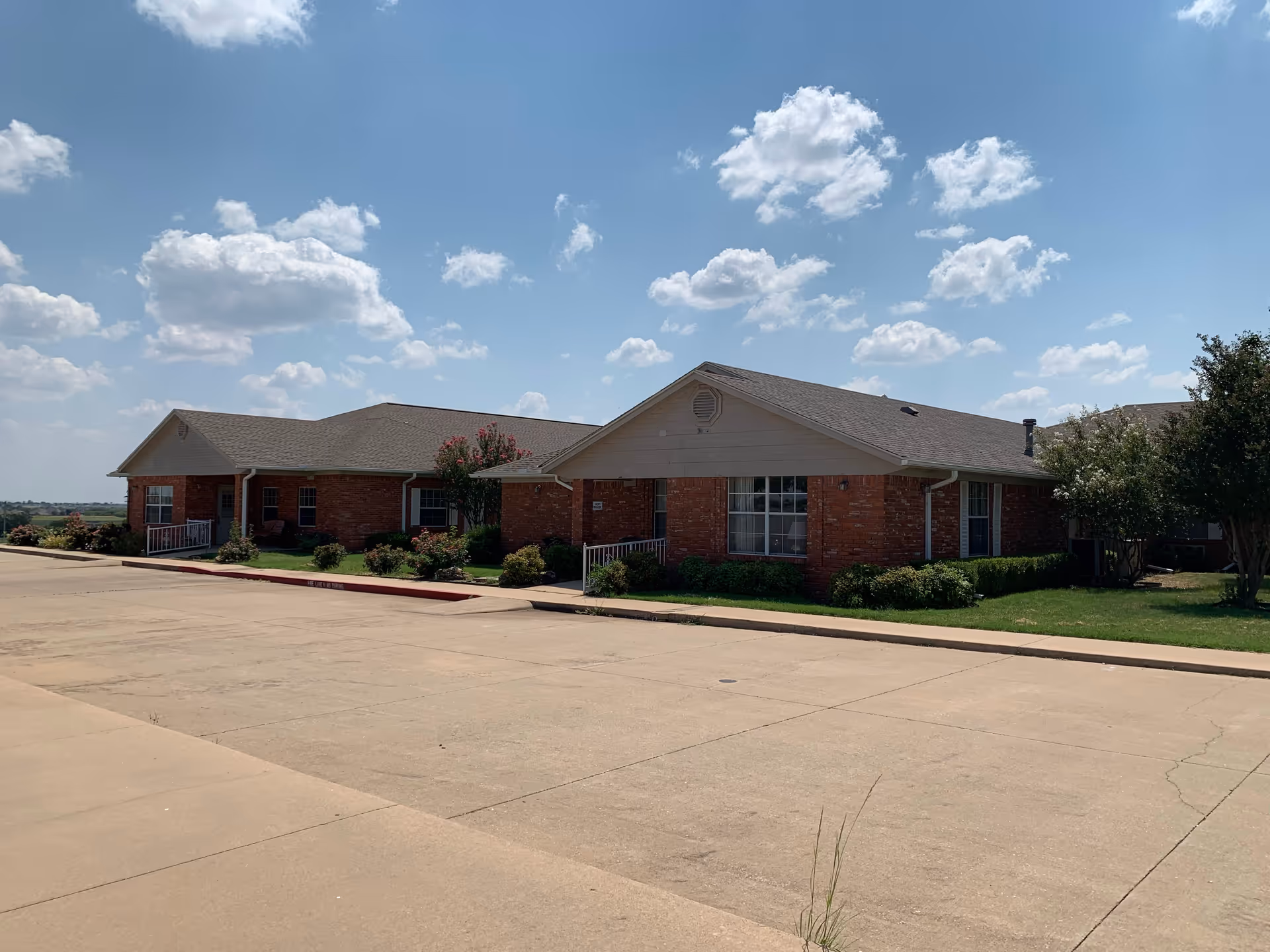 Exterior view of a single-story brick building with a gray roof under a partly cloudy blue sky. The building is surrounded by green shrubs and small trees, with a wide concrete driveway in front.