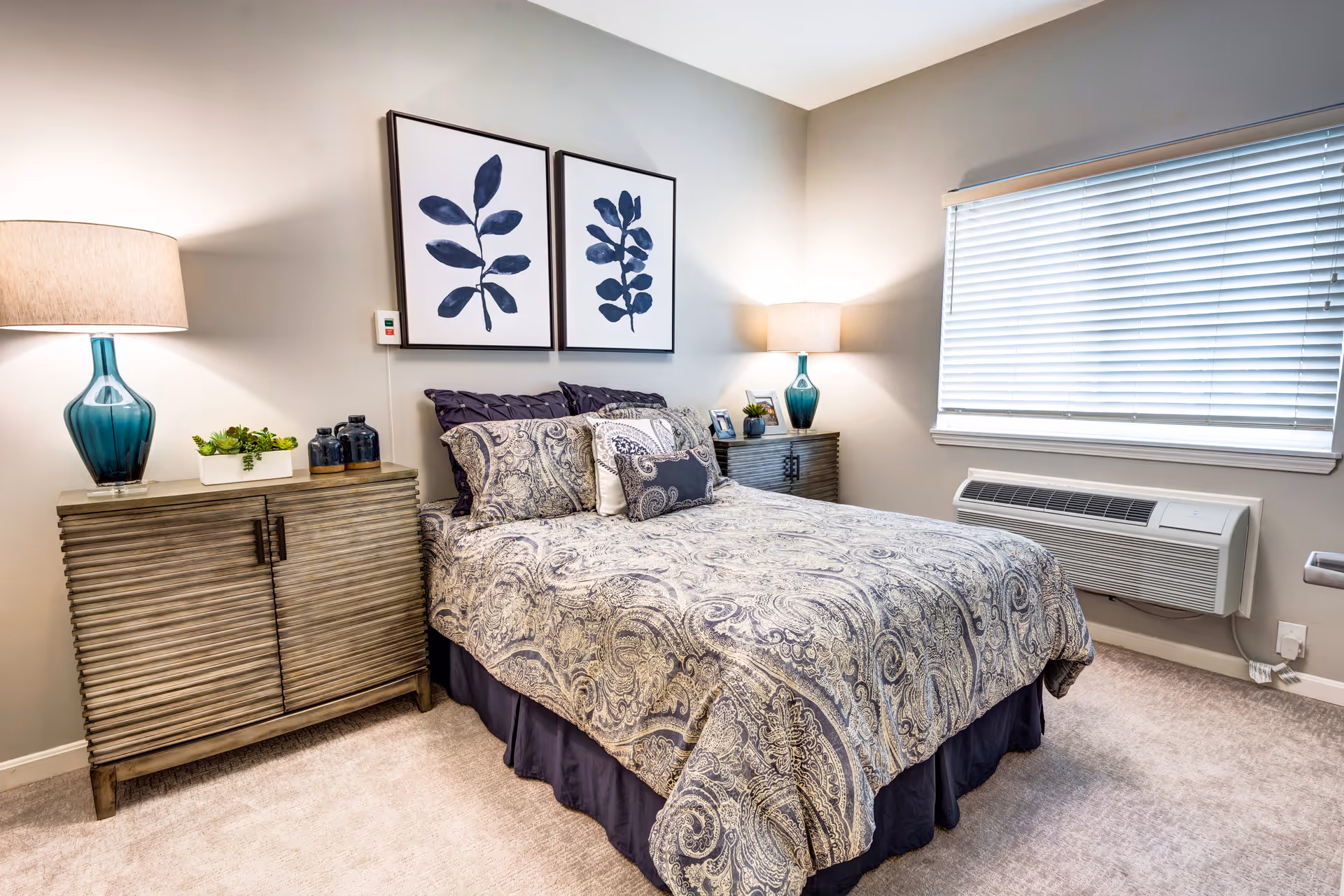 A neatly made bed with patterned bedding and multiple pillows in a bedroom. On either side of the bed are matching wooden nightstands with blue glass lamps and decorative items. Above the bed are two framed botanical prints with blue leaves. A window with closed blinds and a wall-mounted air conditioning unit are visible on the right side of the room.