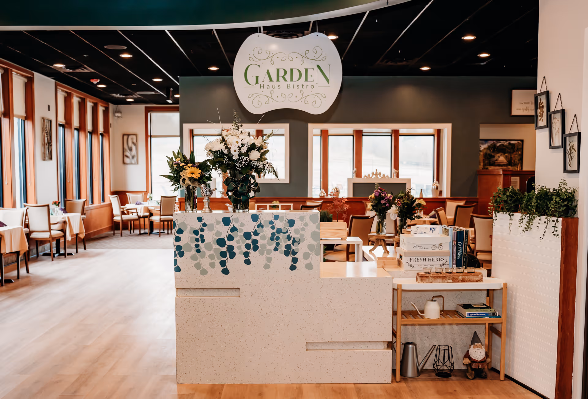 Interior view of a senior living facility dining area named Garden Haus Bistro, featuring a reception desk decorated with floral designs and vases of flowers. The room has wooden floors, multiple tables and chairs arranged for dining, large windows letting in natural light, and decorative plants and framed artwork on the walls.