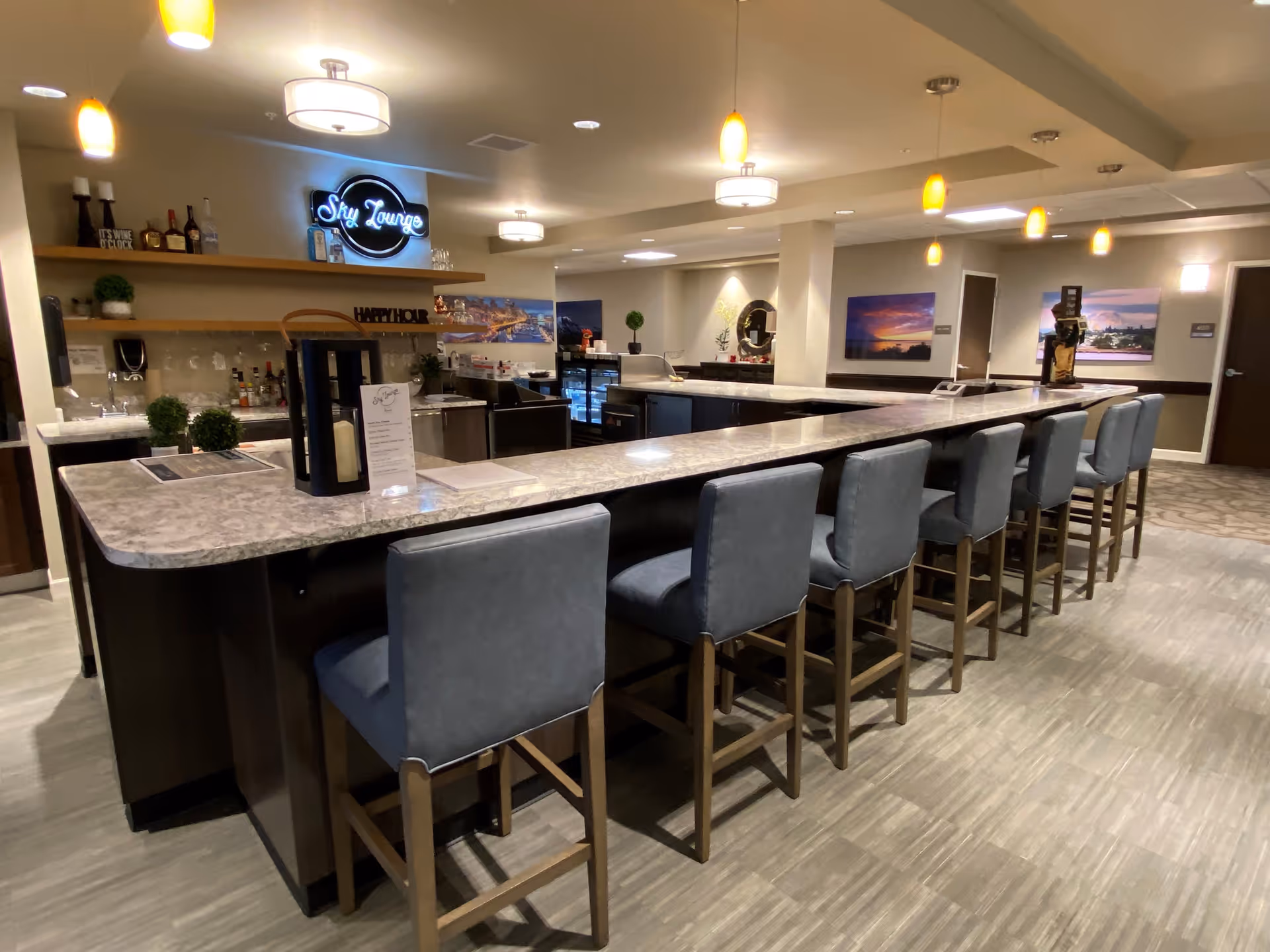 Interior view of a modern lounge area named Sky Lounge with a long marble countertop bar and eight high-back chairs. The bar area has shelves with bottles and decorative items, pendant lights hanging from the ceiling, and framed landscape artwork on the walls.