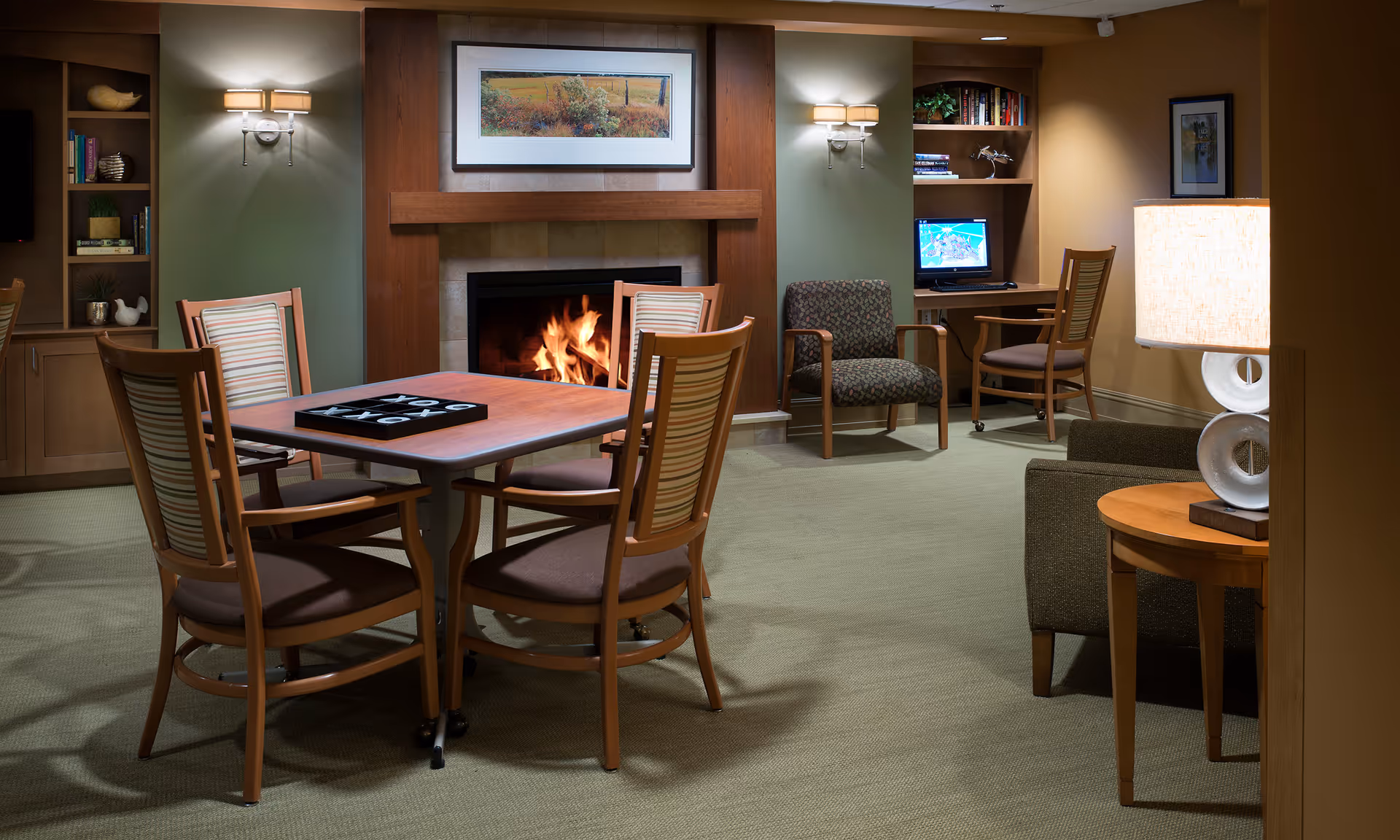 A cozy common area with a wooden table and four chairs in the center, a lit fireplace with a framed landscape painting above it, built-in bookshelves on either side, a computer desk with a chair and monitor, an armchair, and a side table with a lamp.