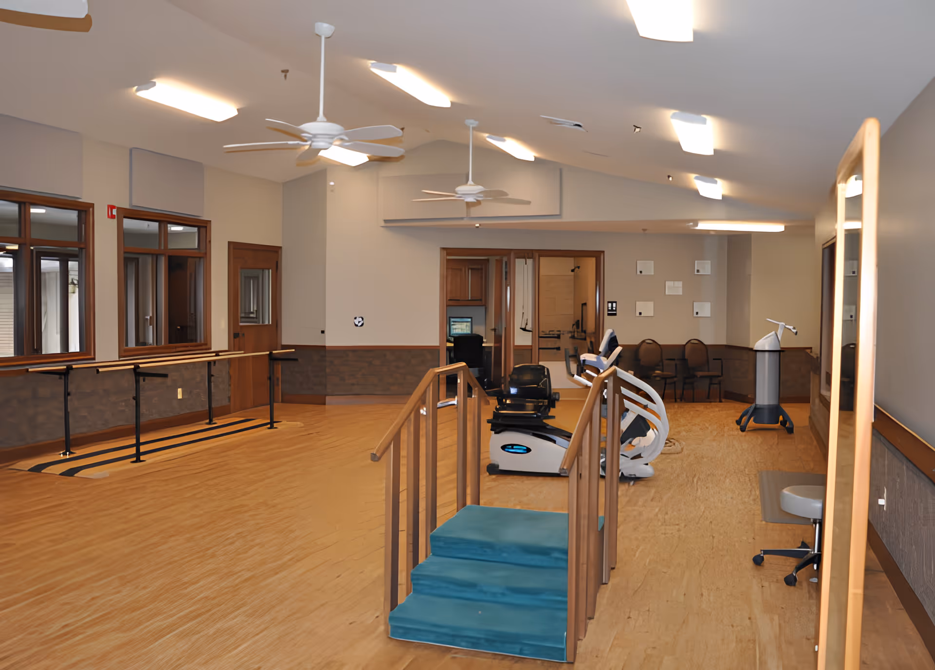A spacious rehabilitation room with wooden flooring, parallel bars for walking exercises, a small set of stairs with handrails, exercise equipment, ceiling fans, and chairs along the back wall. The room has large windows and neutral-colored walls.