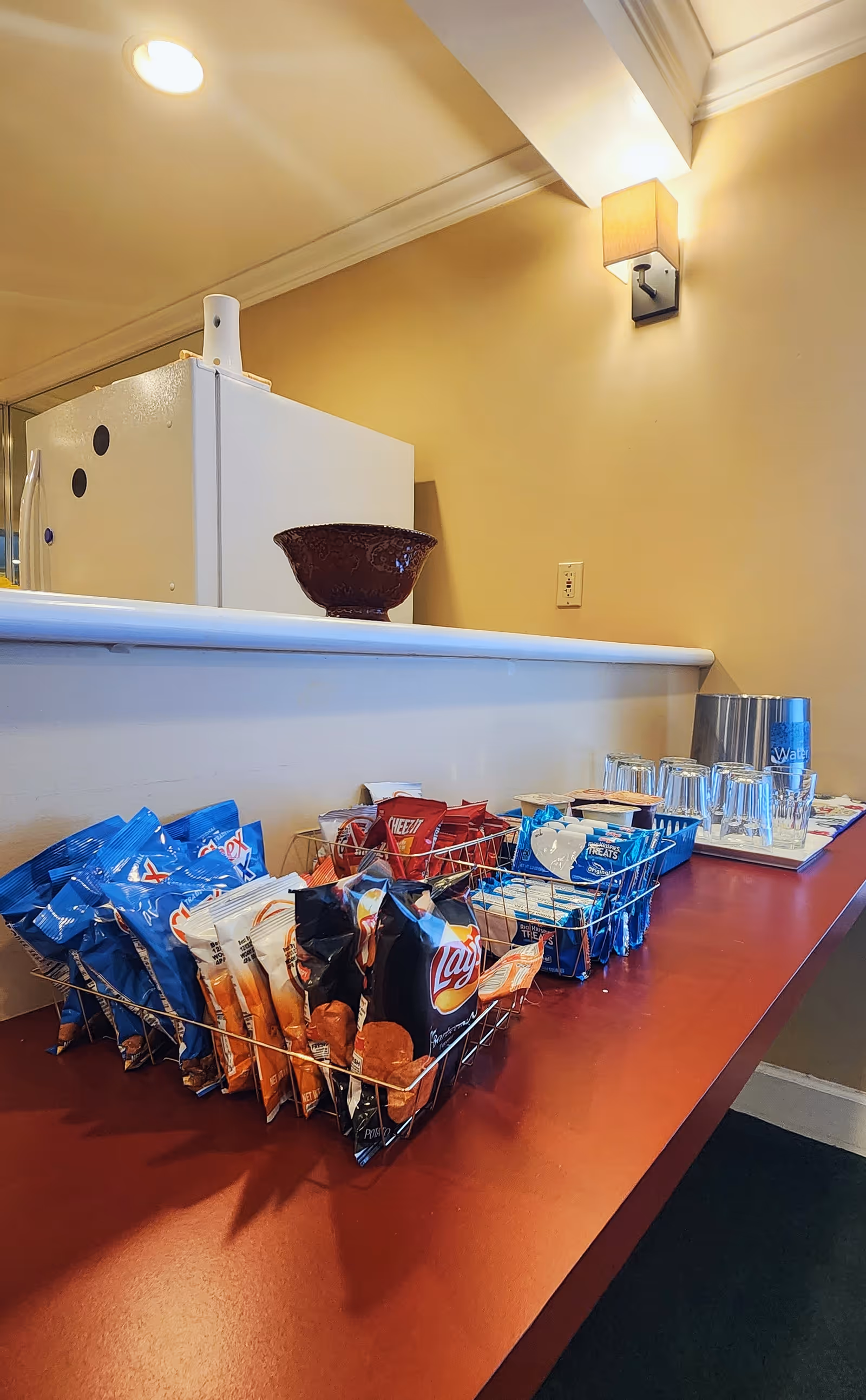 A countertop with baskets filled with various snack bags including Lay's chips, Cheez-It crackers, and other treats. There are also several glasses and a metal container on the right side of the counter. In the background, a white refrigerator and a wall-mounted light fixture are visible.