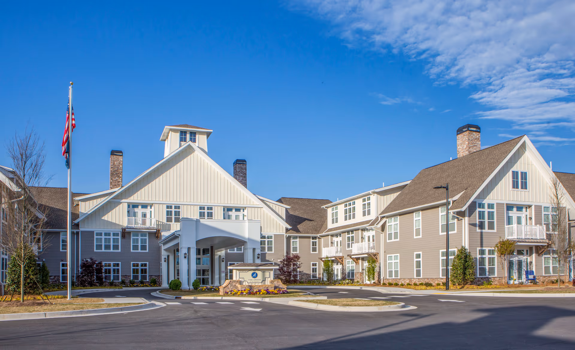 Front exterior view of a large senior living facility building with multiple windows, a covered entrance, two chimneys, and a flagpole with an American flag. The sky is clear and blue.