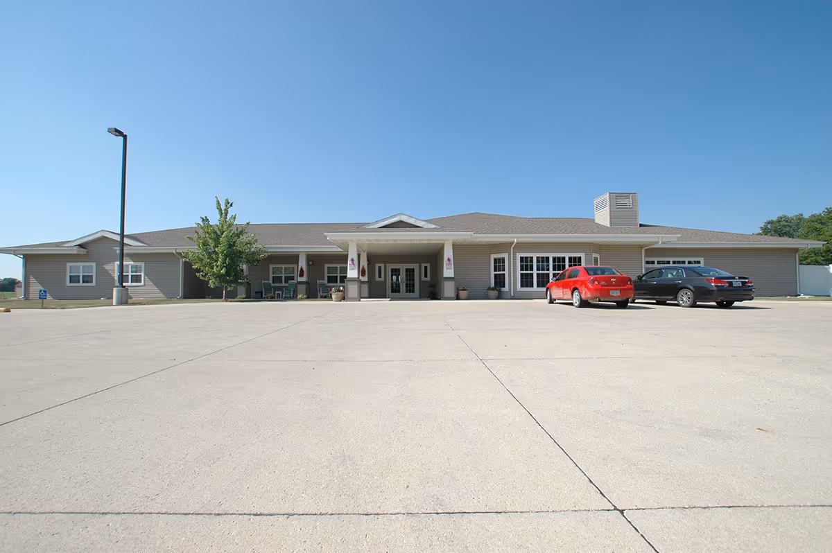 Single-story Windsor Manor building front with a wide concrete parking lot and a few parked cars under a clear blue sky.