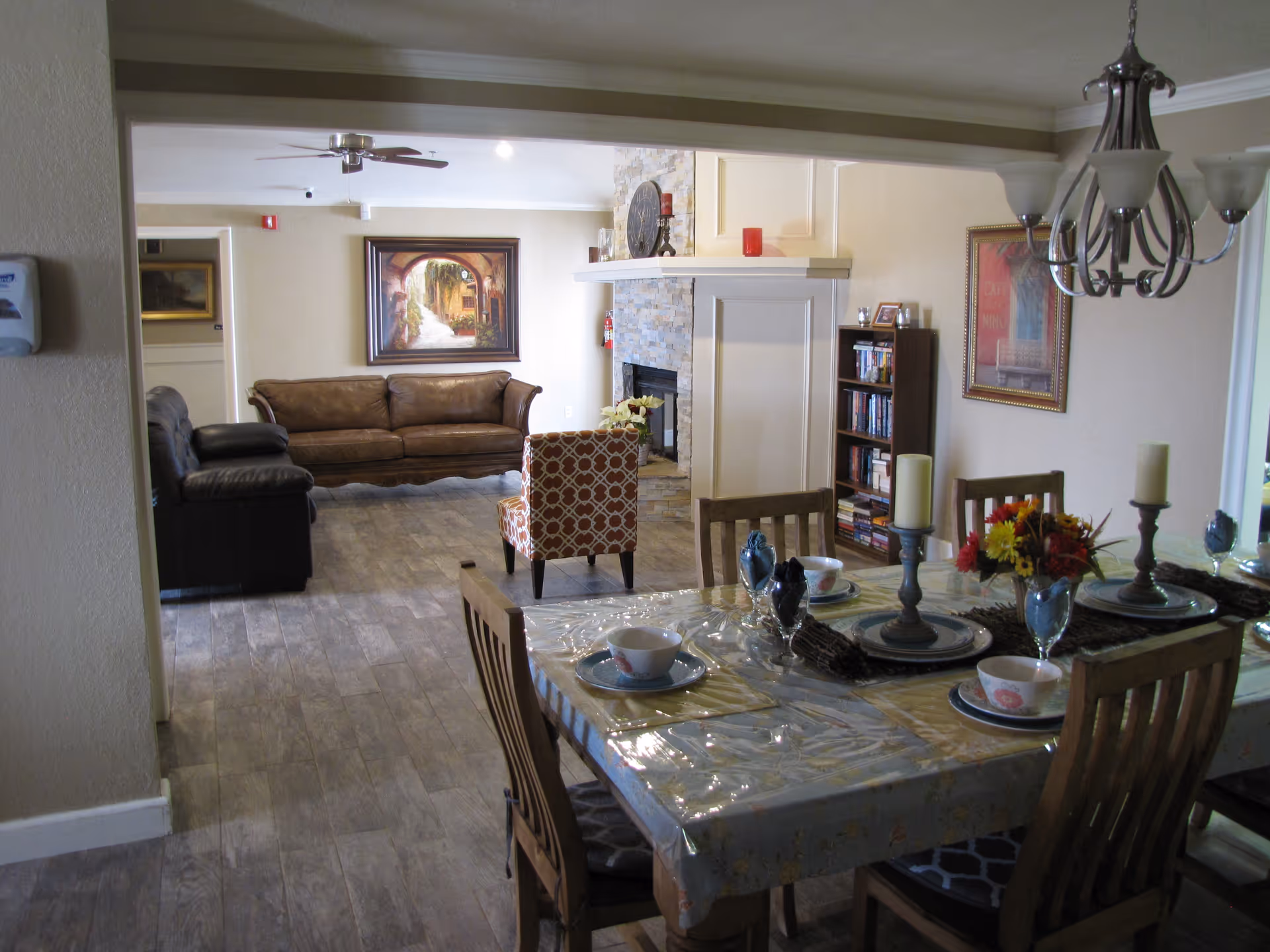 A dining table set for a meal in a communal interior space with chairs, a chandelier, and a living area with couches, a fireplace, and bookshelf visible in the background.
