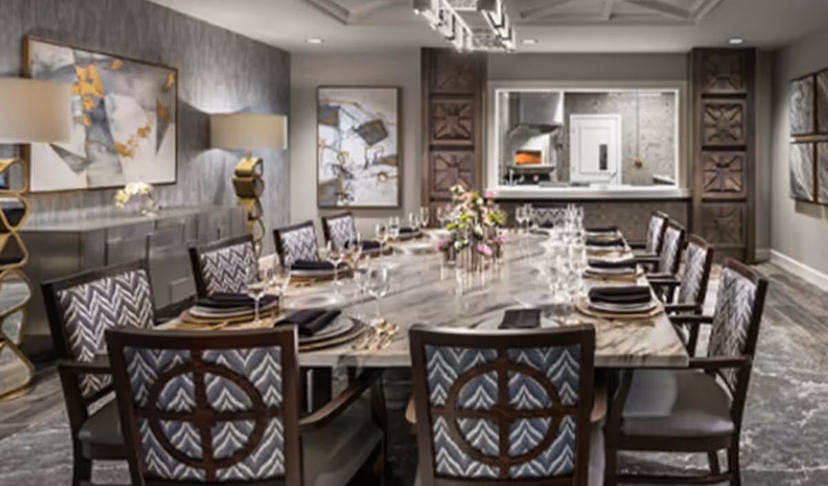 Formal dining room with a long marble table set for a meal, upholstered chairs, artwork on the walls, and a pass-through to a kitchen.