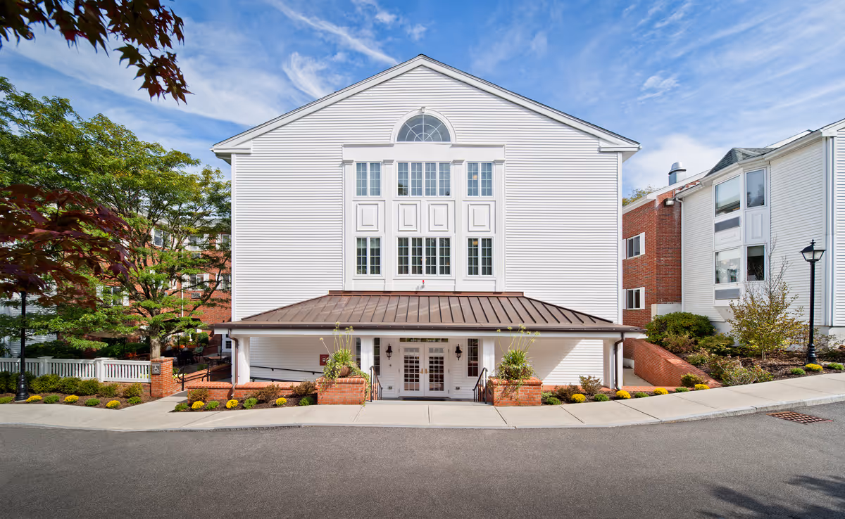 Front view of a white multi-story nursing facility with large windows, a covered entrance, and landscaped beds.