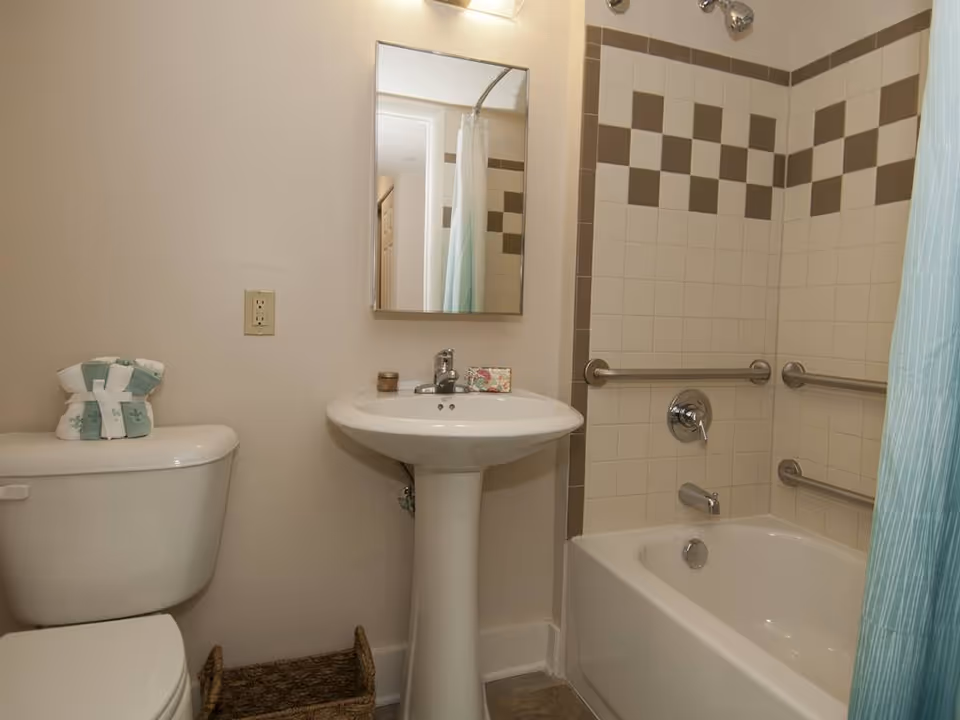 A bathroom with a white toilet, a pedestal sink with a mirror above it, and a bathtub with a shower curtain. The bathtub area has beige and brown checkered tiles and grab bars installed on the walls. There is a small basket on the floor and folded towels on the toilet tank.