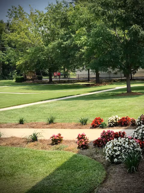 A well-maintained outdoor garden area with green grass, colorful flower beds featuring red, pink, and white flowers, and several large trees providing shade. A paved walkway curves through the garden, and a black metal fence is visible in the background.