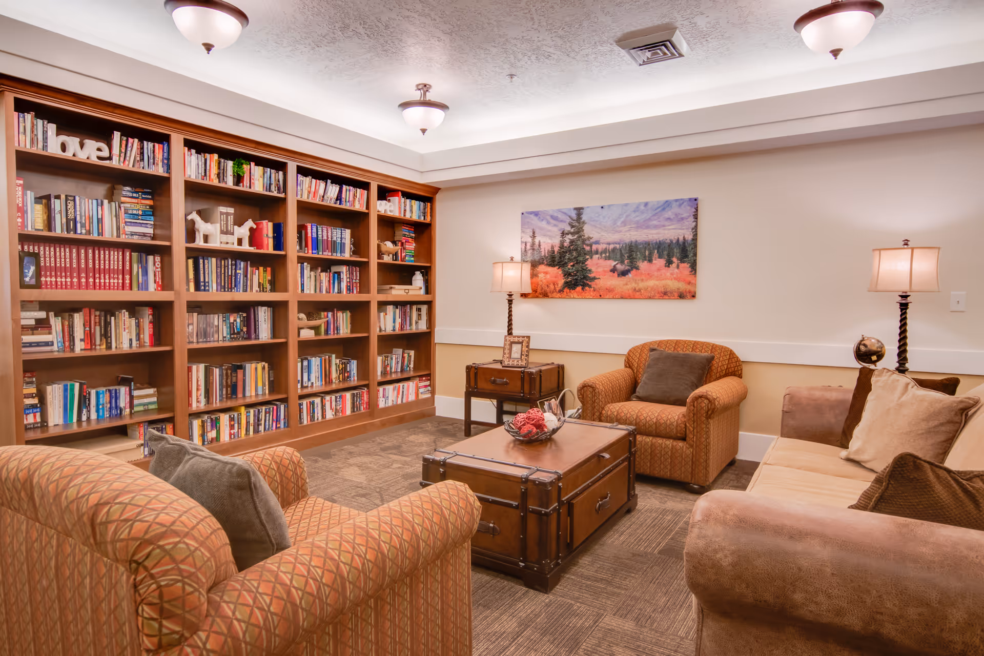 Cozy senior living room with two patterned armchairs, a beige sofa with cushions, a wooden trunk coffee table, and a large bookshelf filled with books. The room is warmly lit with ceiling lights and table lamps, and a landscape painting hangs on the wall.