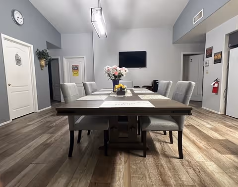 A dining room with a rectangular wooden table surrounded by six upholstered chairs. The table is set with placemats and a centerpiece of flowers. The room has wood flooring, light gray walls, a wall clock, a flat-screen TV mounted on the wall, and several doors leading to other rooms. A fire extinguisher is mounted on the wall near one of the doors.