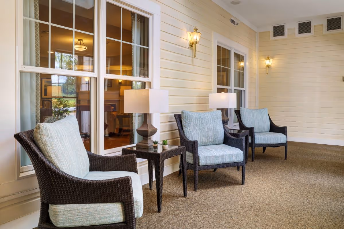 A cozy indoor seating area with three cushioned wicker armchairs arranged along a wall with large windows. Between the chairs are small tables with lamps and small potted plants. The wall is painted light yellow with white trim, and there are two wall-mounted lantern-style lights.
