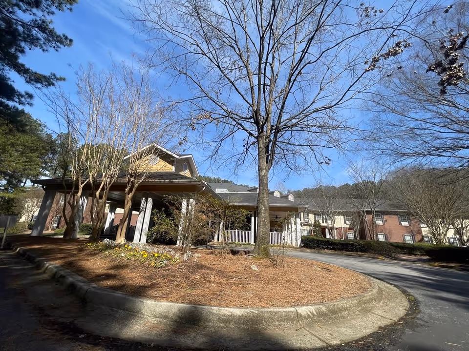 Front entrance and porte-cochère of a senior living building with trees and a circular driveway under a clear blue sky.