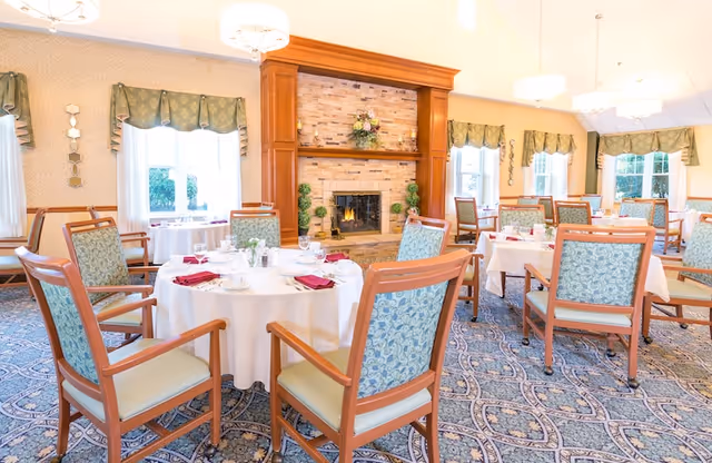 Bright dining room with round tables set for meals, upholstered wooden chairs, and a central fireplace.