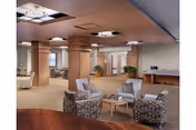 A spacious and well-lit common area in a nursing home featuring comfortable armchairs arranged around small tables. The room has a beige carpet, wooden ceiling panels with recessed lighting, and large columns. There are plants and additional seating visible in the background, creating a welcoming lounge environment.