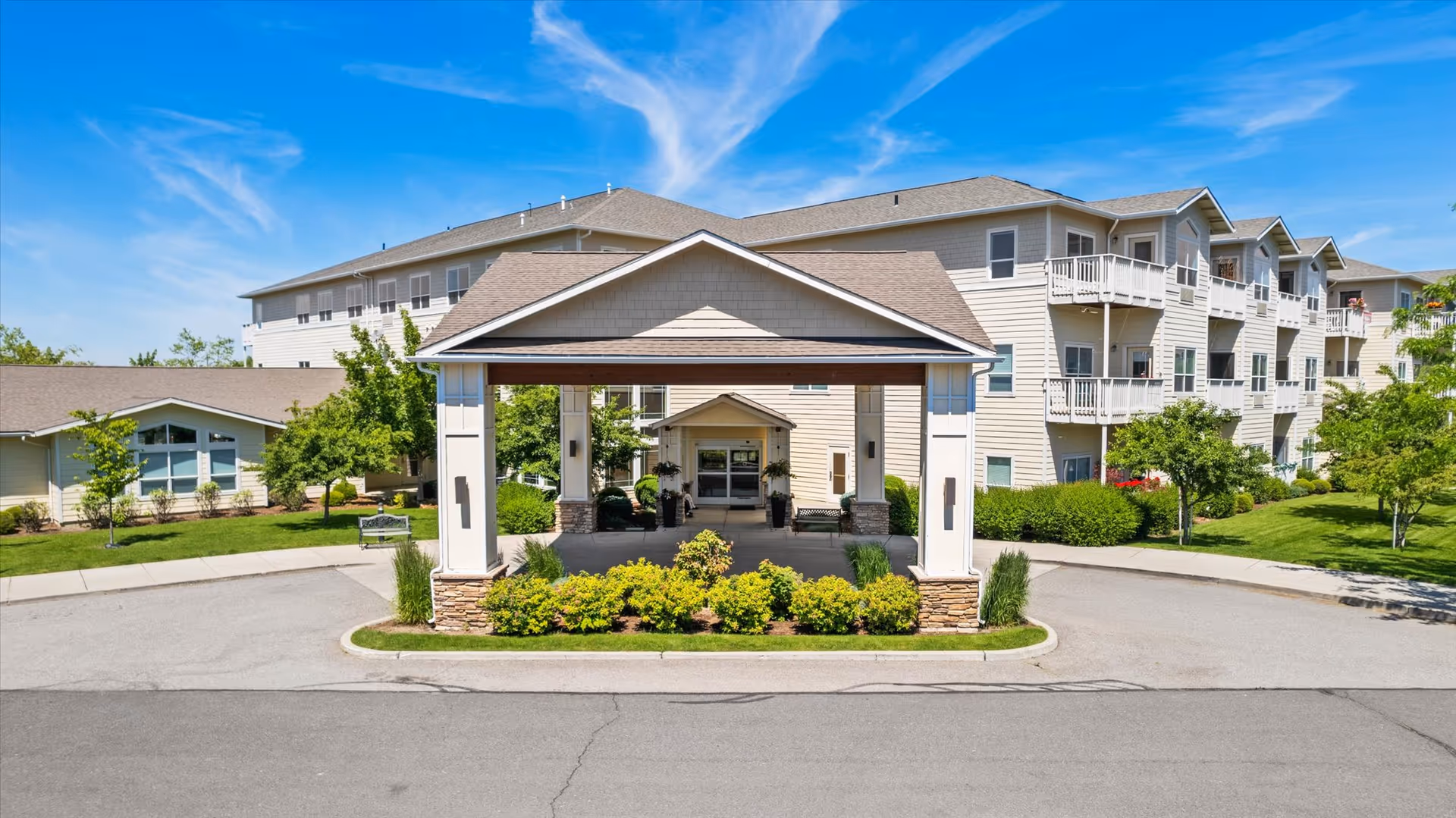 Front exterior view of Vineyard Park of South Hill facility with a covered entrance, surrounded by green shrubs and trees under a blue sky.