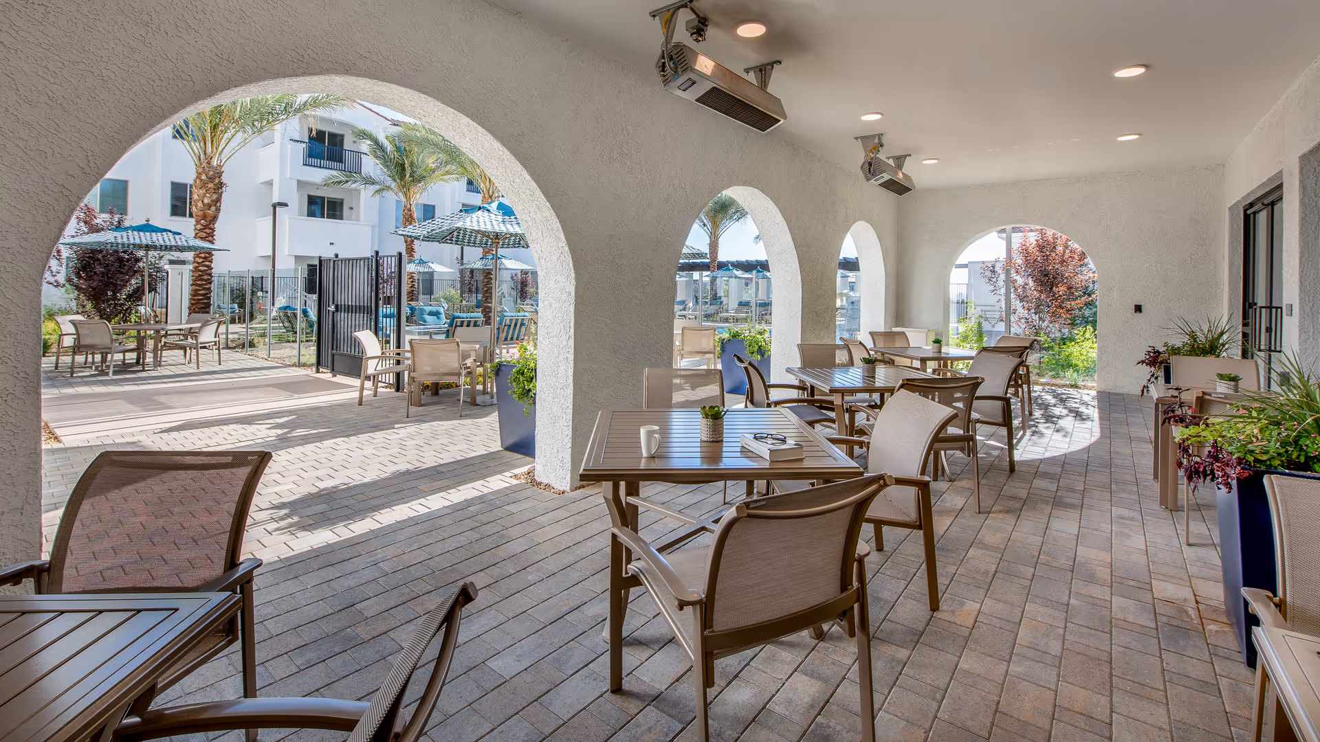 Outdoor covered patio area with multiple tables and chairs arranged for seating. The space features arched openings looking out to a garden area with palm trees, umbrellas, and additional seating. The floor is paved with bricks, and there are potted plants along the sides.
