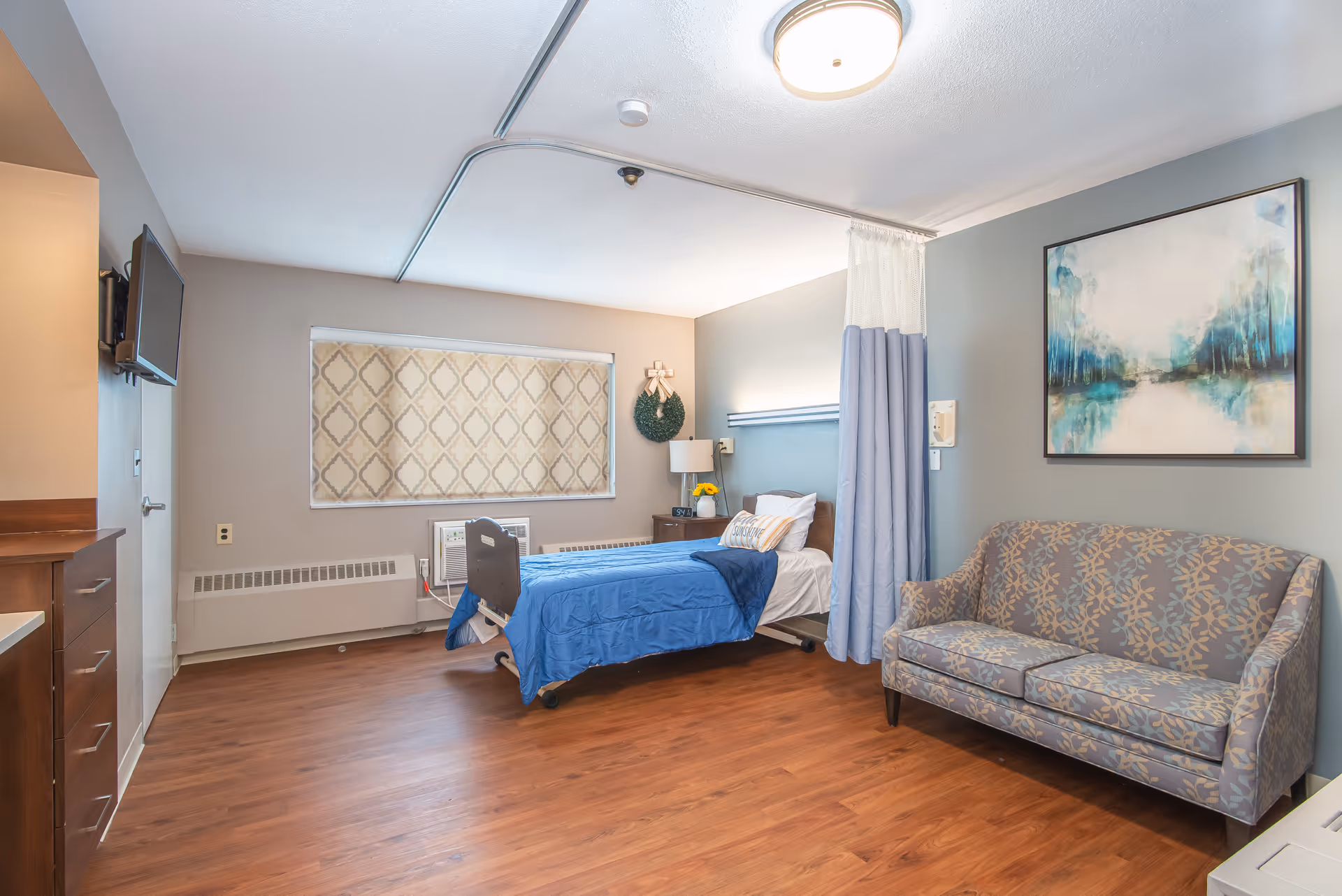 A bright senior-care private room with a hospital-style bed dressed in blue, a patterned sofa, wall art, and wood-look flooring.