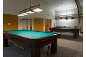Interior view of a recreational room with two billiard tables, overhead lights above each table, wooden doors, and a carpeted floor.