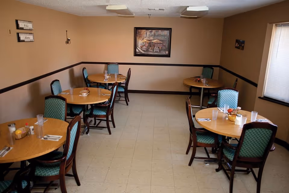 Small dining room with round wooden tables set with glasses and teal upholstered chairs against beige walls.