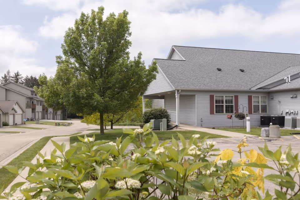 Exterior view of McFarland Villa Assisted Living showing a single-story gray building with red shutters, a large tree, walkway and landscaped shrubs.